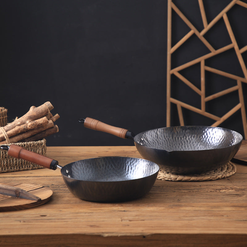 Two black woks with wooden handles on a wooden surface, with a geometric wall art in the background.