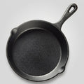 A black cast iron skillet with a long handle, photographed from above on a gray striped cloth over a wooden surface, with cinnamon sticks and star anise nearby.
